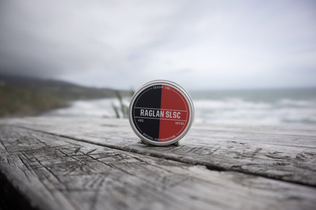 Outdoor Raglan Surf Lifesaving zinc sunscreen on weathered wooden surface with ocean in background, promoting sun protection and safety at Raglan surf beaches in NZ.