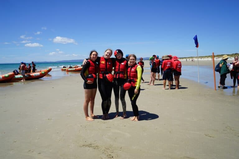 Young female surf lifesavers in red and black uniforms pose with helmets on Raglan beach, showcasing surf rescue skills.